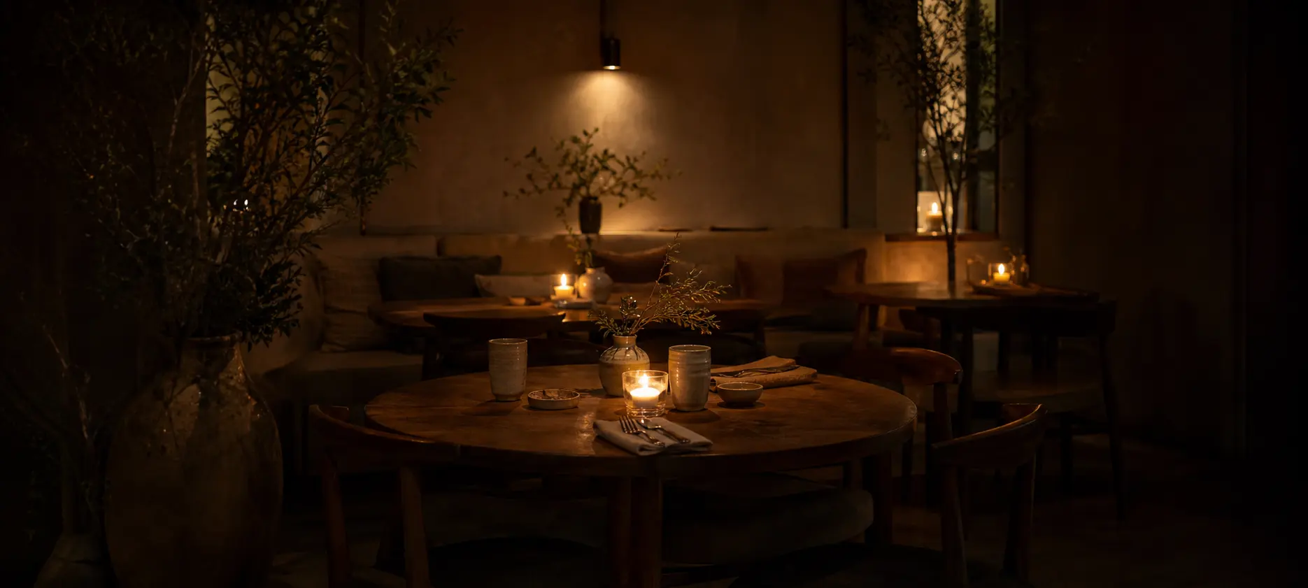 A candlelit corner of the dining room at night: round timber tables and a cushioned banquette lit by clustered candles, with dried branches in the shadows.
