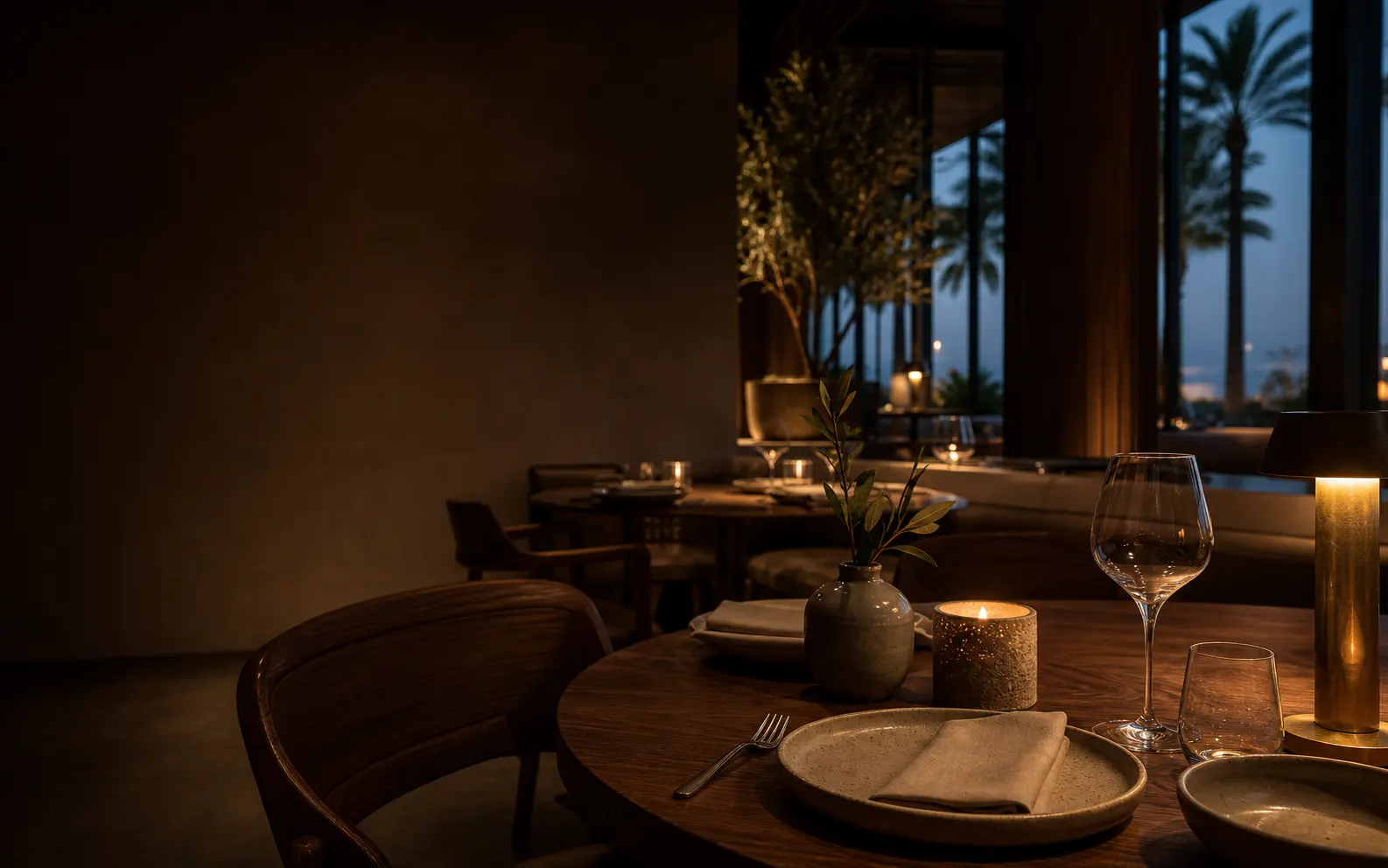A candlelit timber table set for dinner in a warm, dimly lit dining room at dusk, wine glasses and a small vase of greenery, with palm trees silhouetted through the windows beyond.