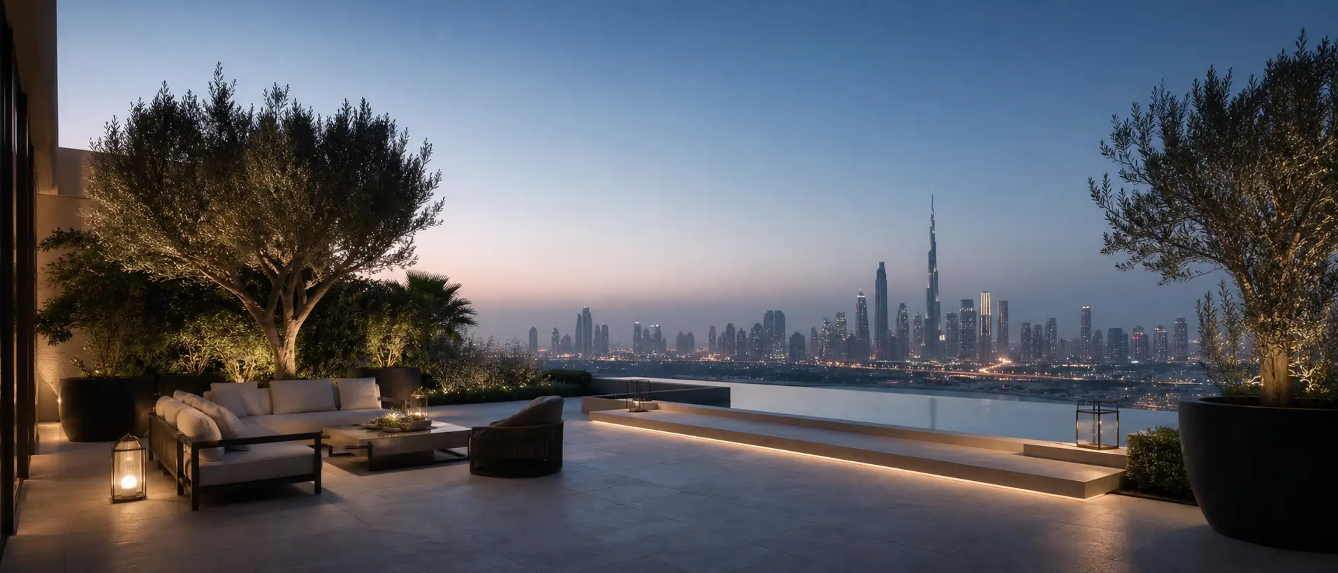 An elevated private terrace at blue hour, a low seating group and olive tree beside an infinity-edge pool that meets the Dubai skyline on the horizon.